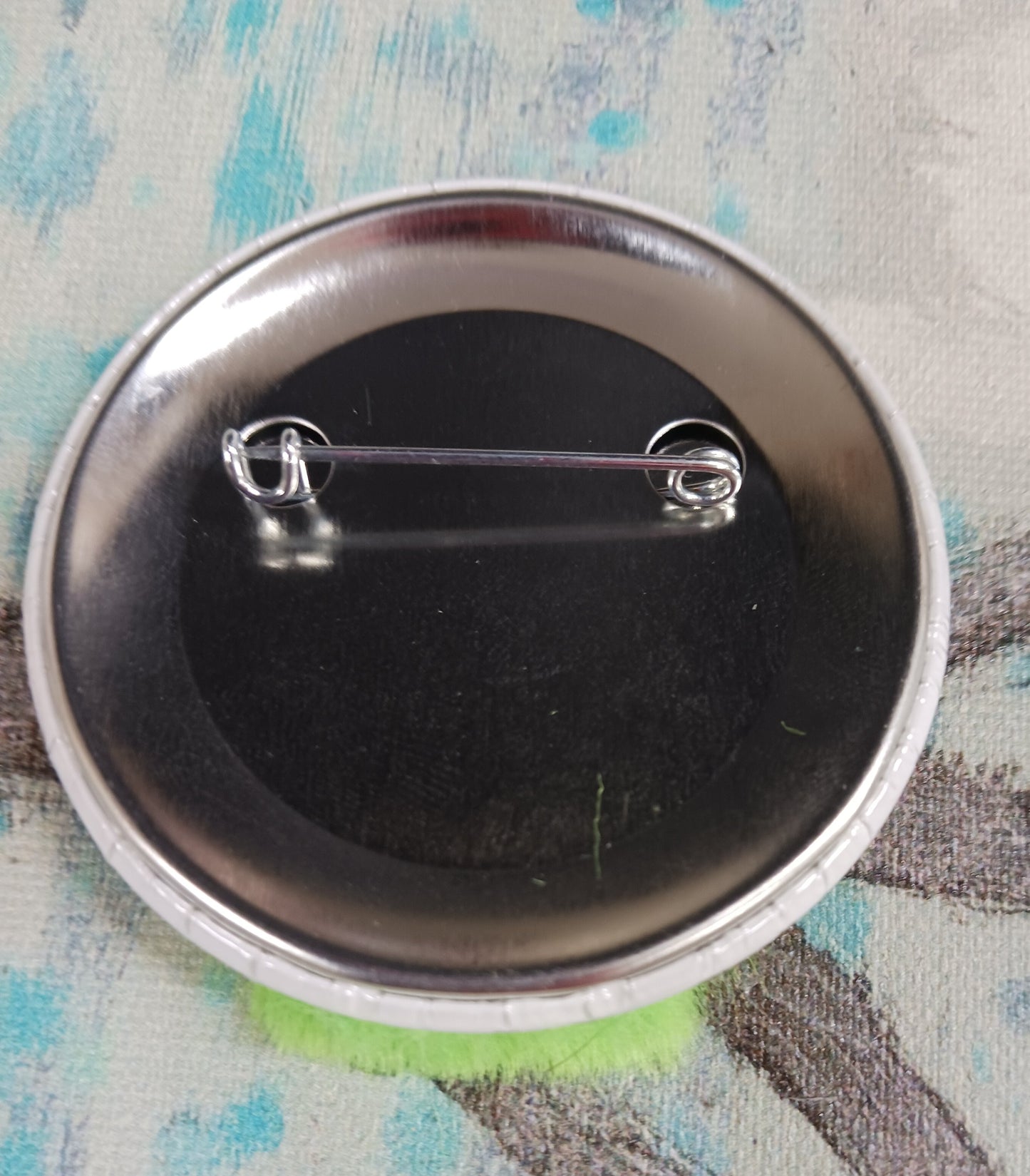 This image shows the back of a pin back button. In this picture you can see the pin that affixes the button to an article of clothing, a backpack, etc.
