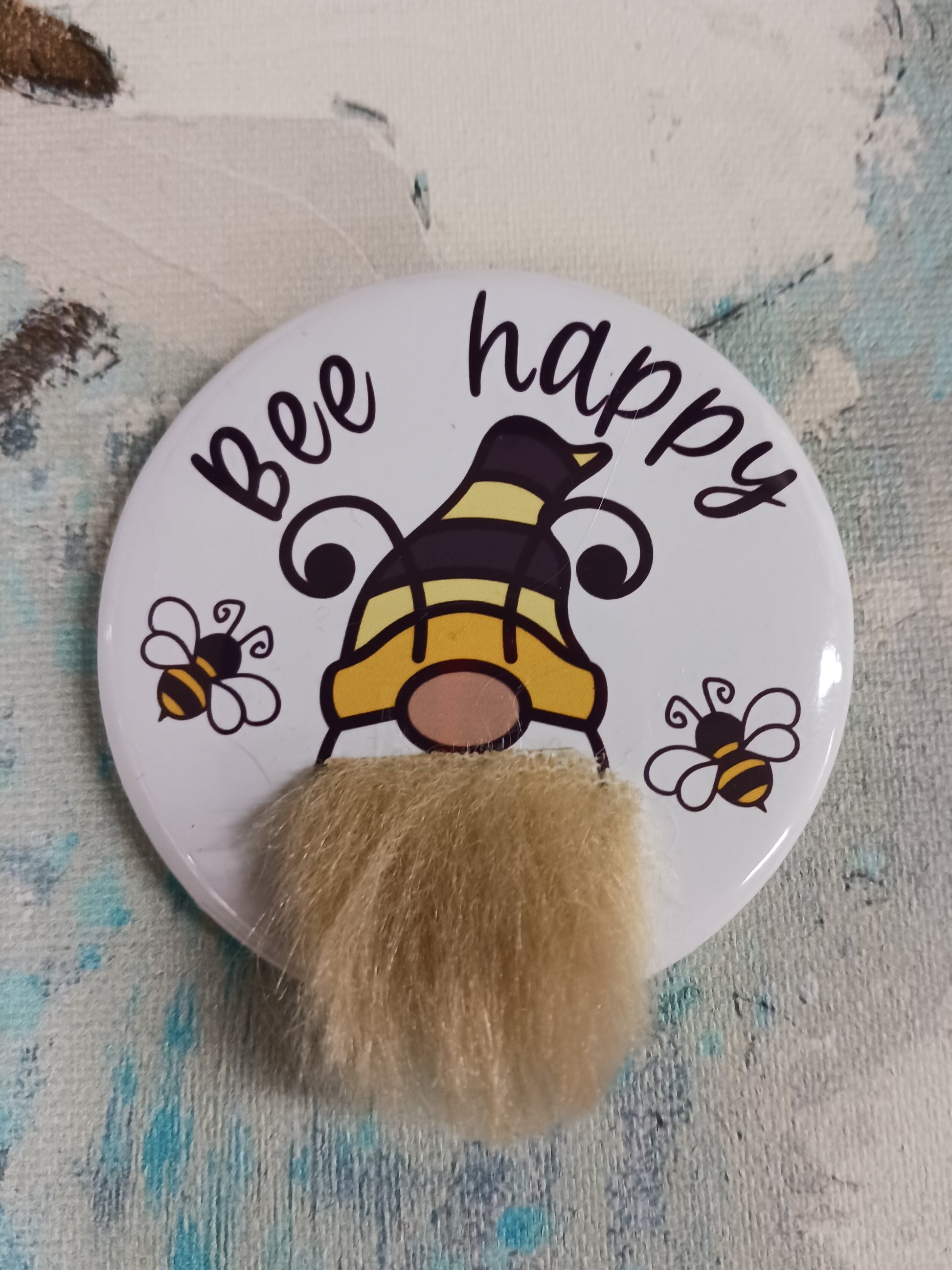 A pin back button that features a bee gnome with a faux fur beard and the phrase Bee Happy appears above his head. He is also accompanied by two small honey bees. 