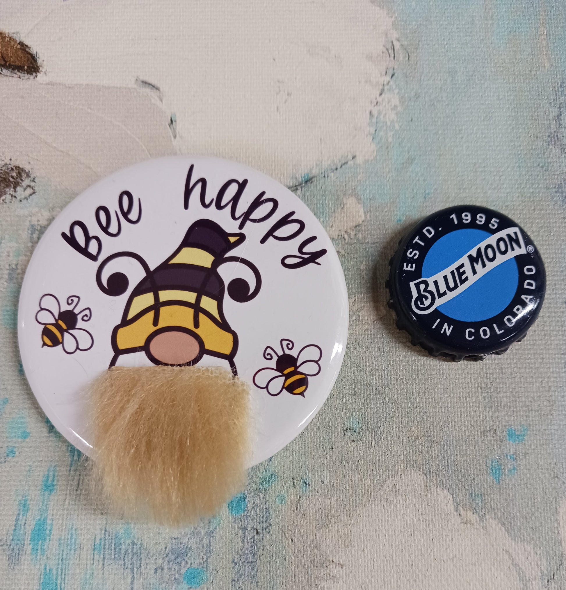 A pin back button that features a bee gnome with a faux fur beard and the phrase Bee Happy appears above his head. He is also accompanied by two small honey bees.  The button is next to a beer bottle cap to show scale. 