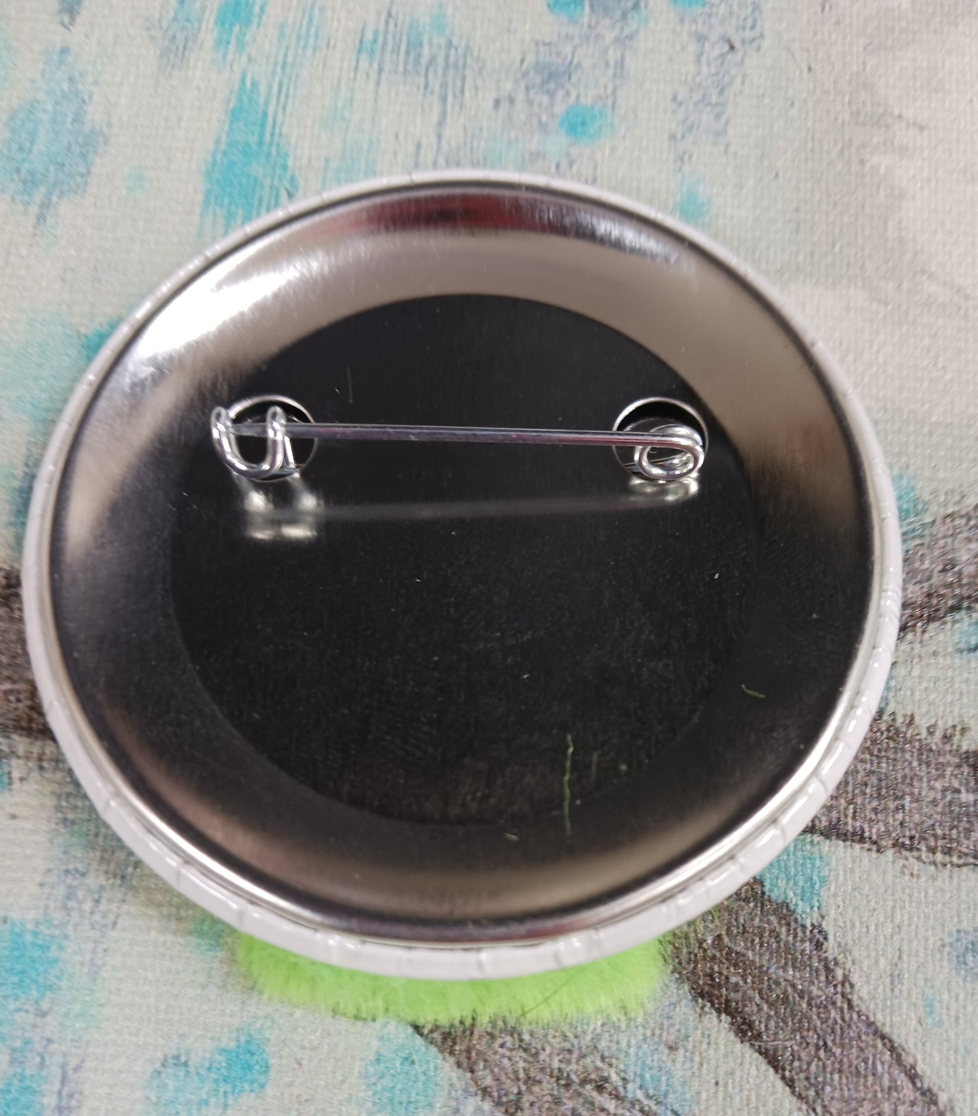 This image shows the back of a pin back button. In this picture you can see the pin that affixes the button to an article of clothing, a backpack, etc.