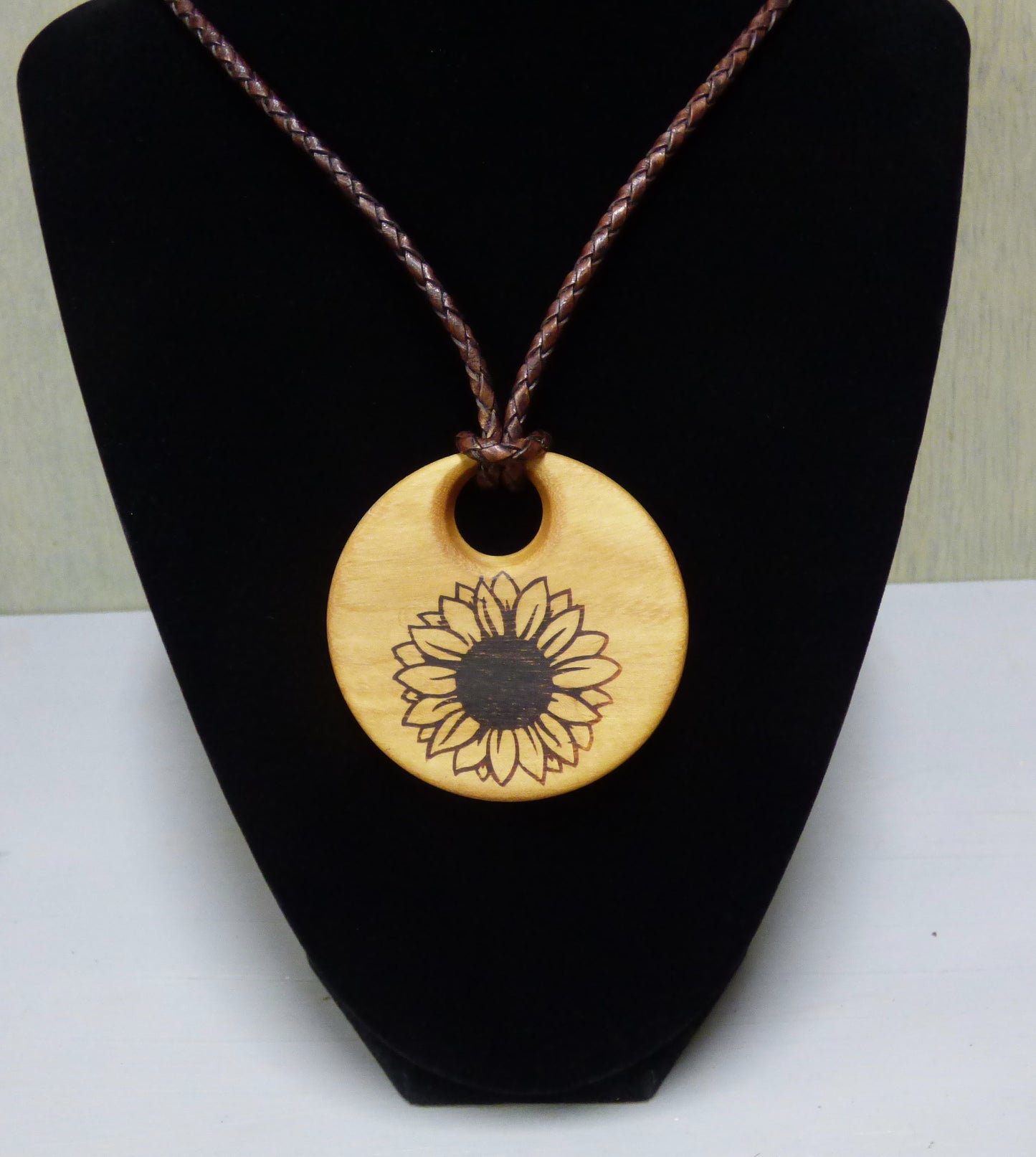 Wooden pendant with sunflower design on a brown cord, displayed on a black stand.