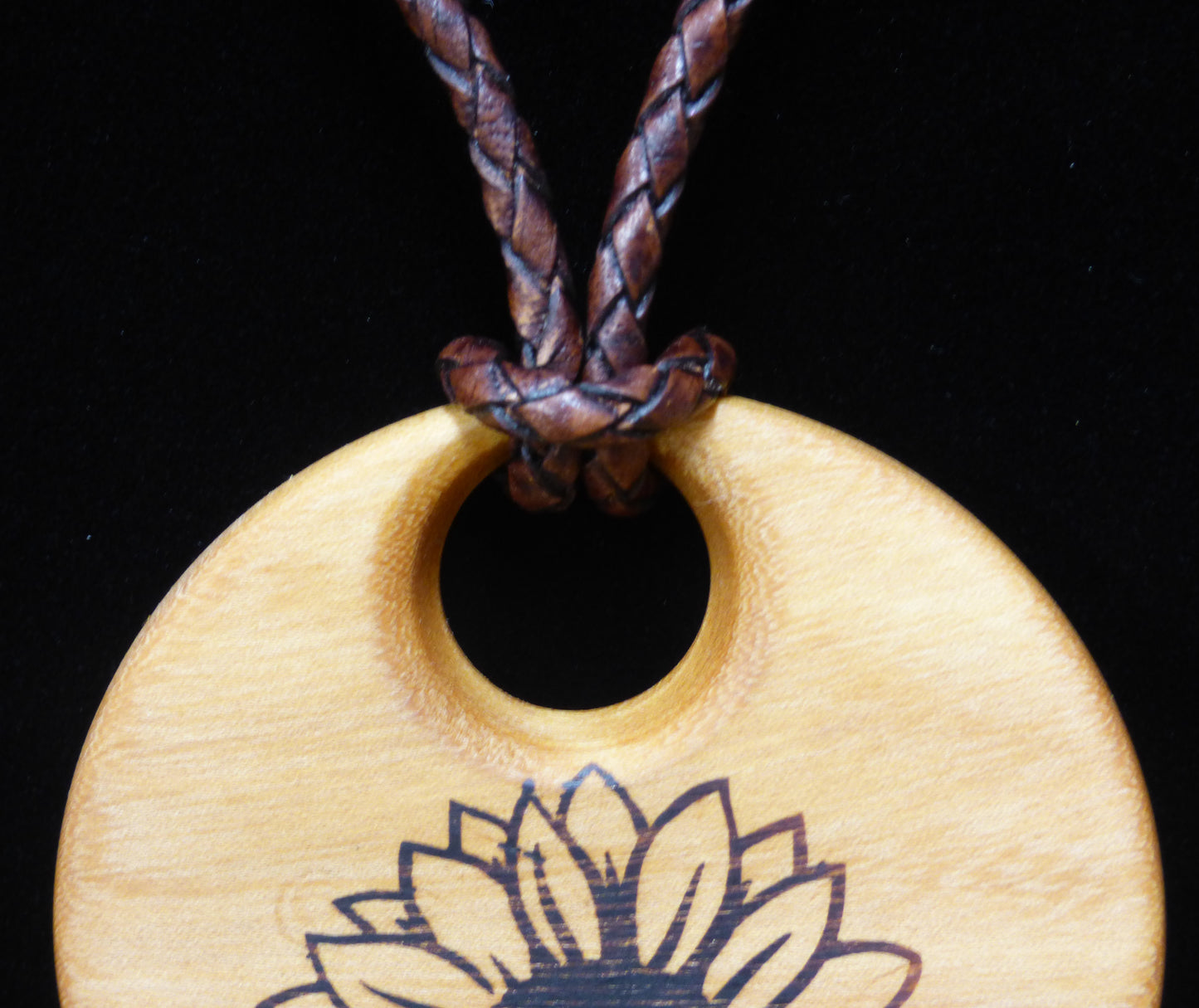 Wooden pendant with sunflower design on a black background with close up of leather cord