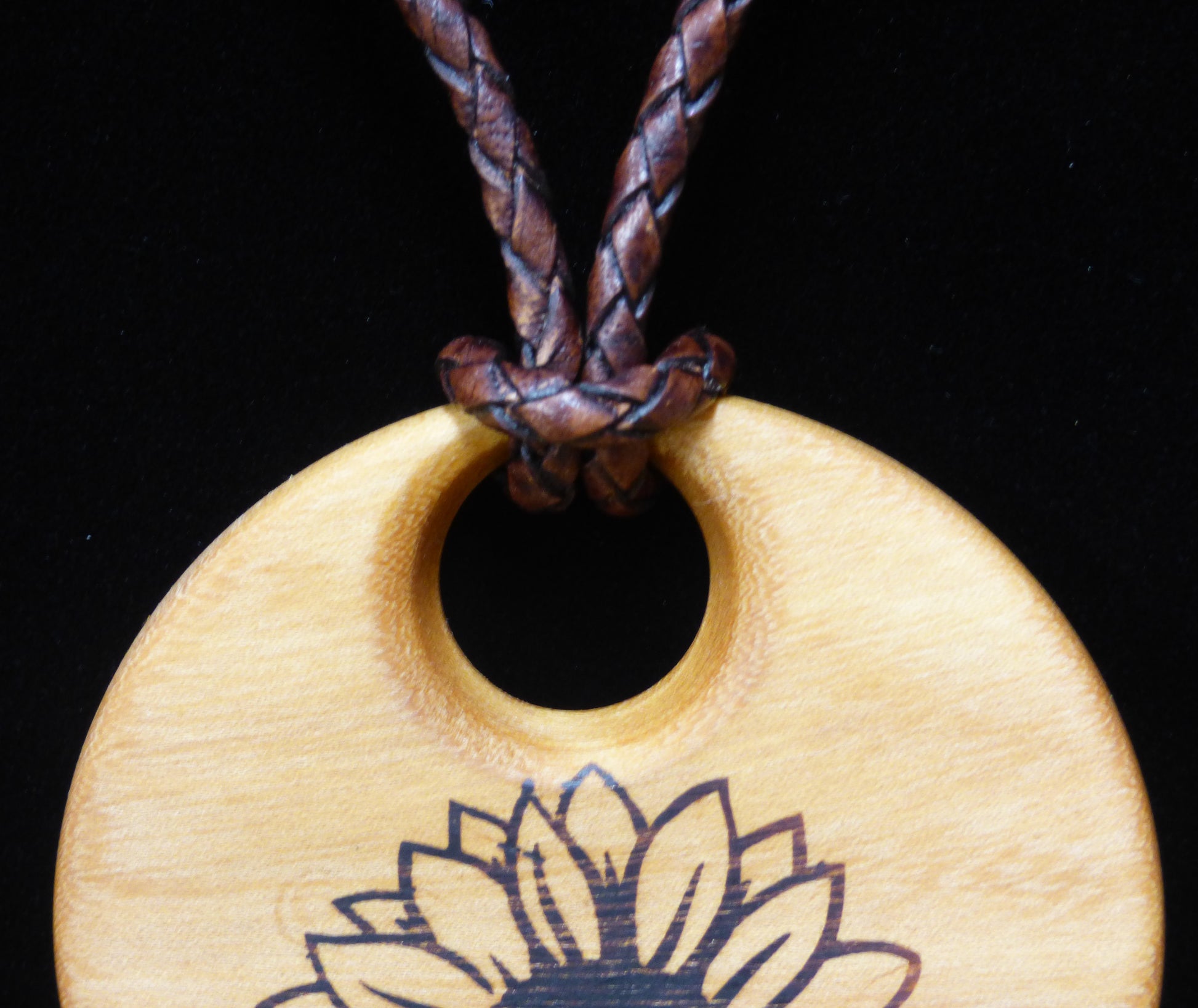 Wooden pendant with sunflower design on a black background with close up of leather cord