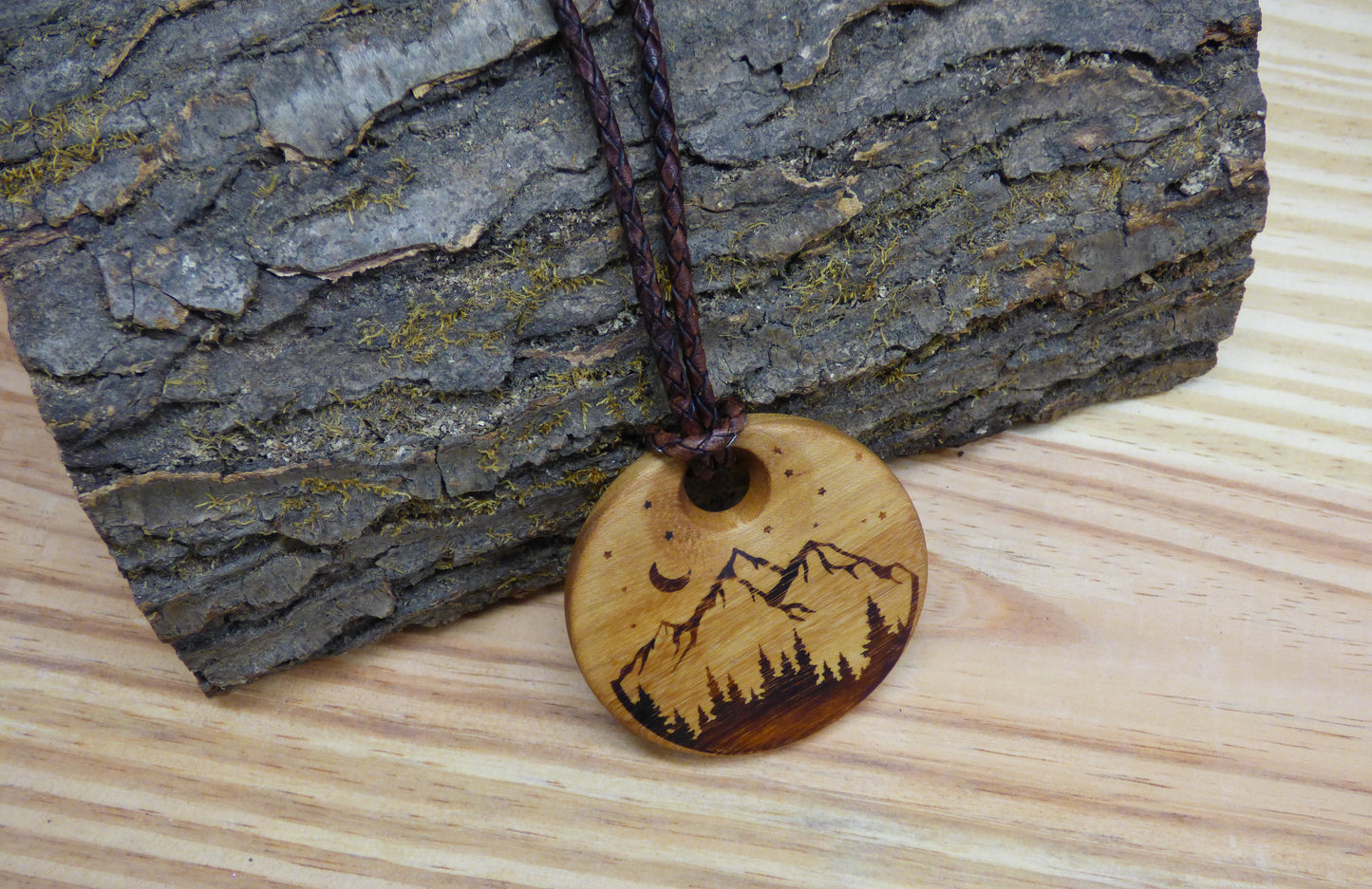 Wooden pendant with moon, stars, and mountain design with braided leather cord against a wood log and wood surface background